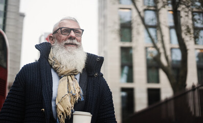 Hipster senior business man drinking coffee while walking to office with city in background - Focus...