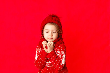 a little girl in winter clothes makes a wish with her eyes closed on a red background. New year's concept