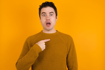 Young handsome Caucasian man wearing yellow sweater against orange wall being in stupor shocked, has astonished expression pointing at oneself with finger saying: Who me?