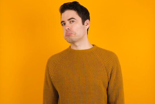 Young handsome Caucasian man wearing yellow sweater against orange wall with snobbish expression curving lips and raising eyebrows, looking with doubtful and skeptical expression, suspect and doubt.