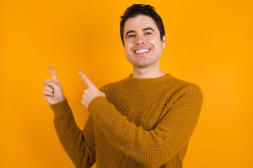 Young handsome Caucasian man wearing yellow sweater against orange wall with positive expression, indicates with fore finger at blank copy space for your promotional text or advertisement.