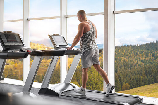 Muscular athletic bodybuilder fitness model running treadmill gym near big window