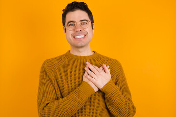 Honest Young handsome Caucasian man wearing yellow sweater against orange wall keeps hands on...