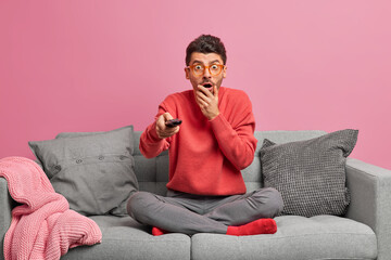 Impressed stunned adult man stares at camera watches horrible movie holds remote control sits in...