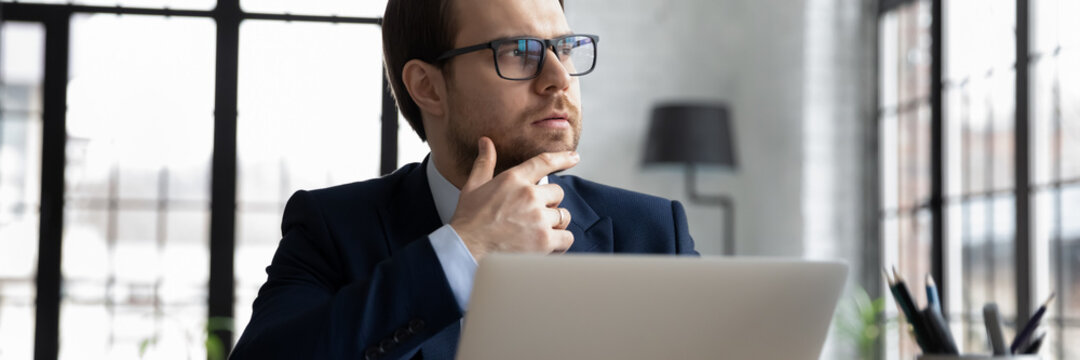 Close up thoughtful motivated businessman wearing glasses looking to aside in office horizontal banner, touching chin, sitting with laptop, pensive entrepreneur planning, pondering business strategy