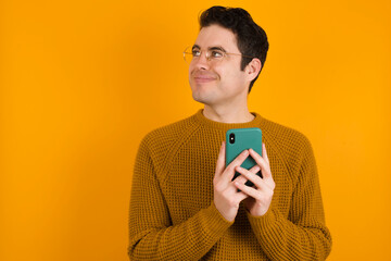 Happy Young handsome Caucasian man wearing yellow sweater against orange wall listening to music with earphones using mobile phone.
