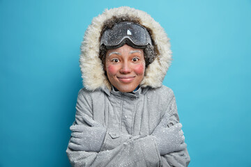 Winter holidays and recreation concept. Cheerful frozen woman feels cold after going snowboarding in mountains trembles and hugs herself to warm wears grey jacket with hood gloves feels satisfied - obrazy, fototapety, plakaty