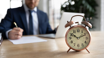 Close up alarm clock on desk, businessman wearing suit using laptop, working with correspondence on...