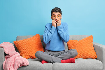 Shocked Caucasian man watches horror film reacts on scarying scene sits crossed legs on sofa in...