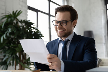 Close up smiling satisfied businessman wearing glasses reading letter, sitting at desk in office, working with correspondence, excited employee received job promotion, business achievement