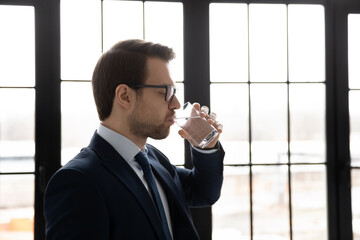 Side view confident businessman wearing glasses drinking fresh pure mineral water, standing near...
