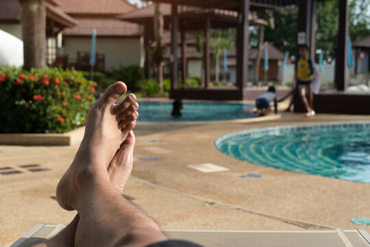Feet of the man are crossed. Lie down on a hammock By the pool. Blurred background image behind swimming pool with children in the resort.