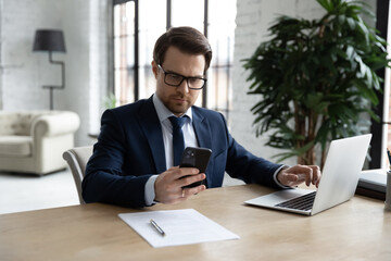 Obraz premium Confident businessman wearing glasses using phone and laptop, working on online project, statistics, sitting at desk in modern office, serious employee looking at smartphone screen, reading email