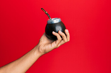 Hand of hispanic man holding cup of mate infusion beverage over isolated red background.