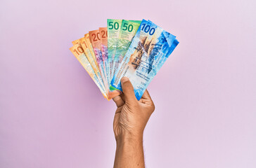 Hispanic hand holding swiss franc banknotes over isolated pink background.