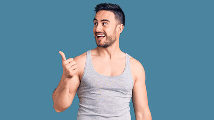 Young handsome man wearing swimwear and sleeveless t-shirt smiling with happy face looking and pointing to the side with thumb up.
