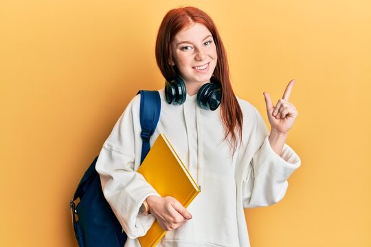 Young red head girl wearing student backpack and holding book smiling happy pointing with hand and finger to the side