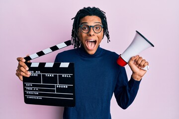 Young african american man holding video film clapboard and megaphone celebrating crazy and amazed...