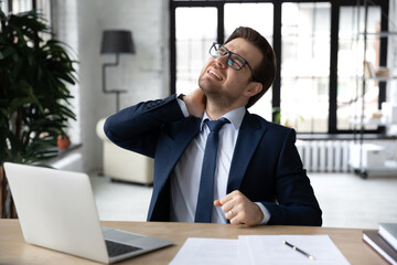 Unhappy young businessman wearing glasses massaging stiff neck muscles, sitting at work desk in office, feeling pain after long sedentary work, exhausted employee feeling unhealthy and unwell
