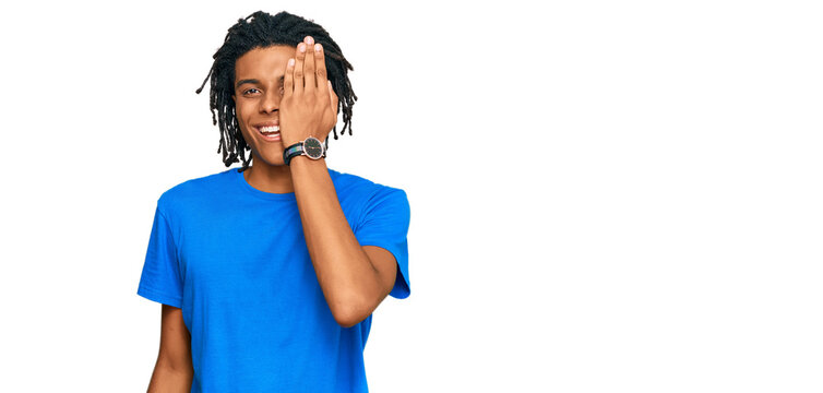 Young african american man wearing casual clothes covering one eye with hand, confident smile on face and surprise emotion.