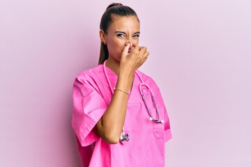 Young hispanic woman wearing doctor uniform and stethoscope smelling something stinky and...