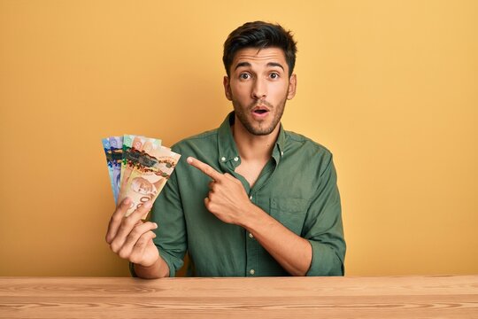 Young handsome man holding canadian dollars surprised pointing with finger to the side, open mouth amazed expression.