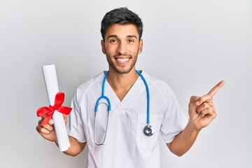 Young handsome man wearing doctor uniform holding medical degree smiling happy pointing with hand and finger to the side