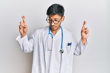 Young handsome man wearing doctor uniform and stethoscope gesturing finger crossed smiling with hope and eyes closed. luck and superstitious concept.