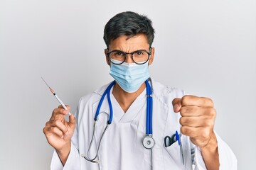 Young handsome man wearing doctor uniform and medical mask holding syringe annoyed and frustrated shouting with anger, yelling crazy with anger and hand raised