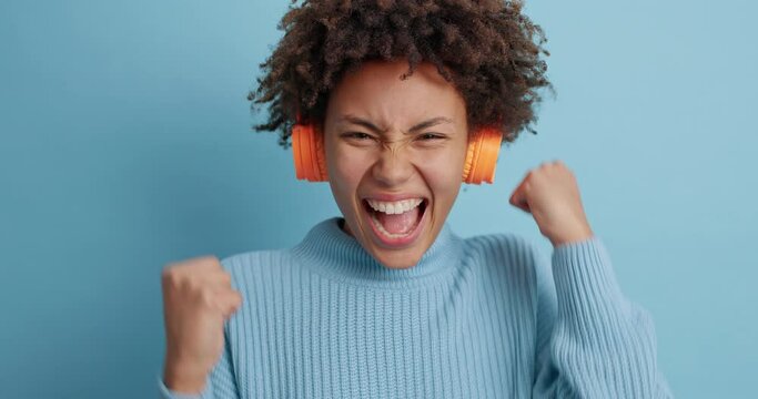 Stunned wondered woman stares surprisingly at camera reacts on something awesome drops jaw makes yes gesture raises clenched fists wears wireless headphones on ears isolated over blue background