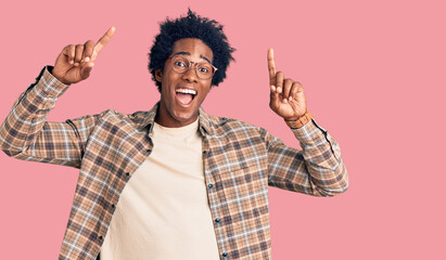 Handsome african american man with afro hair wearing casual clothes and glasses smiling amazed and surprised and pointing up with fingers and raised arms.