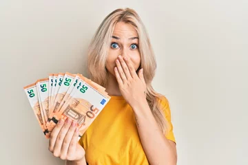 Fotobehang Beautiful caucasian blonde girl holding bunch of 50 euro banknotes covering mouth with hand, shocked and afraid for mistake. surprised expression © Krakenimages.com