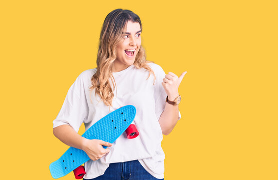 Young caucasian woman holding skate pointing thumb up to the side smiling happy with open mouth