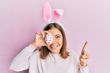 Obraz na płótnie Canvas Young beautiful woman wearing cute easter bunny ears holding egg smiling with an idea or question pointing finger with happy face, number one
