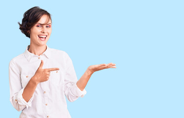 Beautiful young woman with short hair wearing elegant white shirt amazed and smiling to the camera while presenting with hand and pointing with finger.