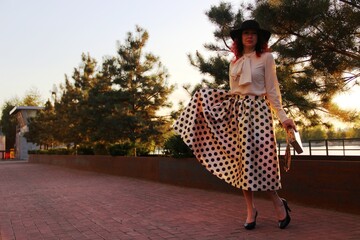 portrait in sunset. a woman in a black hat, beige blouse and a midi skirt with black polka dots in the bright rays of the sunset walks in a city park on the embankment. horizontal photo