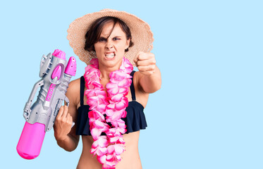 Beautiful young woman with short hair wearing bikini and hawaiian lei holding water gun annoyed and frustrated shouting with anger, yelling crazy with anger and hand raised