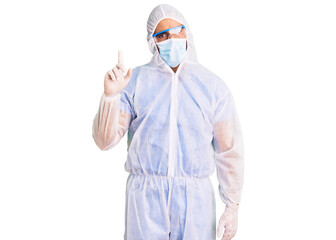 Young hispanic man wearing doctor protection coronavirus uniform and medical mask showing and pointing up with finger number one while smiling confident and happy.