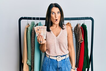 Middle age brunette personal shopper woman holding 10 pounds banknotes scared and amazed with open...