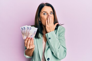 Middle age brunette woman holding russian 500 ruble banknotes covering mouth with hand, shocked and...