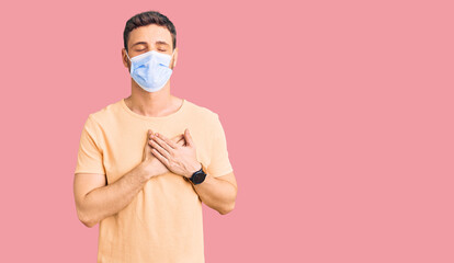 Handsome young man with bear wearing medical mask for coronavirus smiling with hands on chest with closed eyes and grateful gesture on face. health concept.