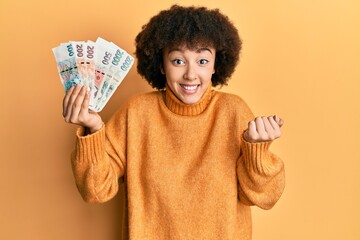 Young hispanic girl holding czech koruna banknotes screaming proud, celebrating victory and success very excited with raised arm