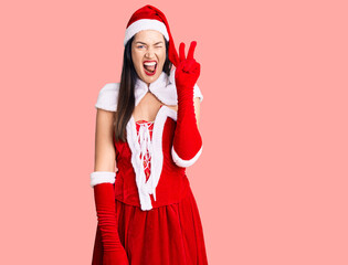 Young beautiful caucasian woman wearing santa claus costume smiling with happy face winking at the camera doing victory sign with fingers. number two.