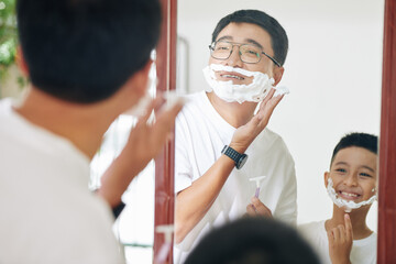 Smiling mature man and his teenage son looking at mirror and applying shaving cream
