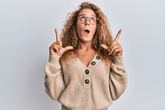 Beautiful caucasian teenager girl wearing casual clothes and glasses amazed and surprised looking up and pointing with fingers and raised arms.
