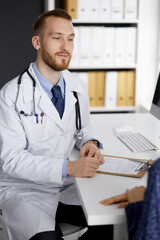 Friendly red-bearded doctor and patient woman discussing current health examination while sitting in sunny clinic. Medicine concept