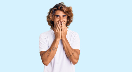 Young hispanic man wearing casual white tshirt laughing and embarrassed giggle covering mouth with...