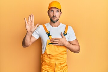 Young handsome man wearing handyman uniform over yellow background swearing with hand on chest and...