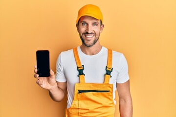 Young handsome man wearing handyman uniform showing smartphone screen looking positive and happy standing and smiling with a confident smile showing teeth