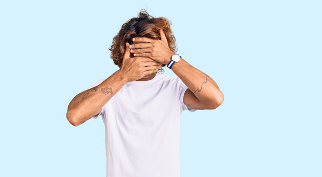 Young hispanic man wearing casual white tshirt covering eyes and mouth with hands, surprised and shocked. hiding emotion
